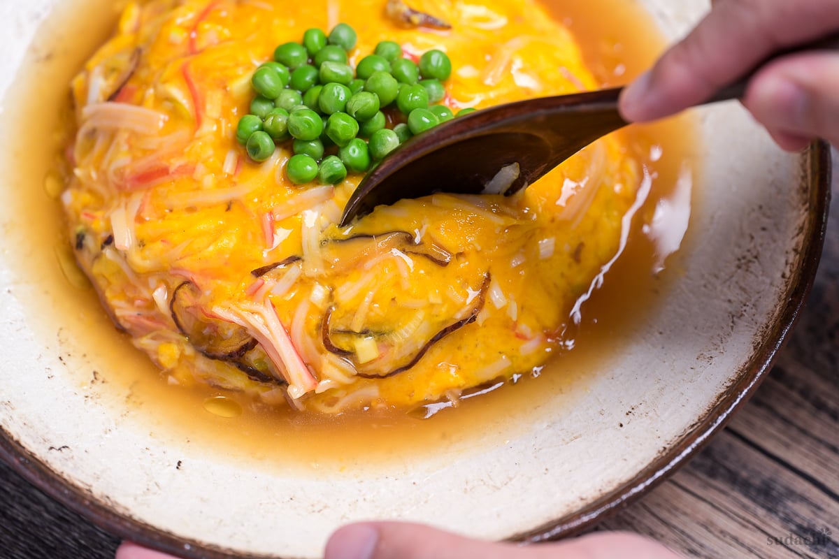 A spoon digging into tenshinhan (Japanese crab omelette with rice) topped with green peas on a textured cream plate with brown rim
