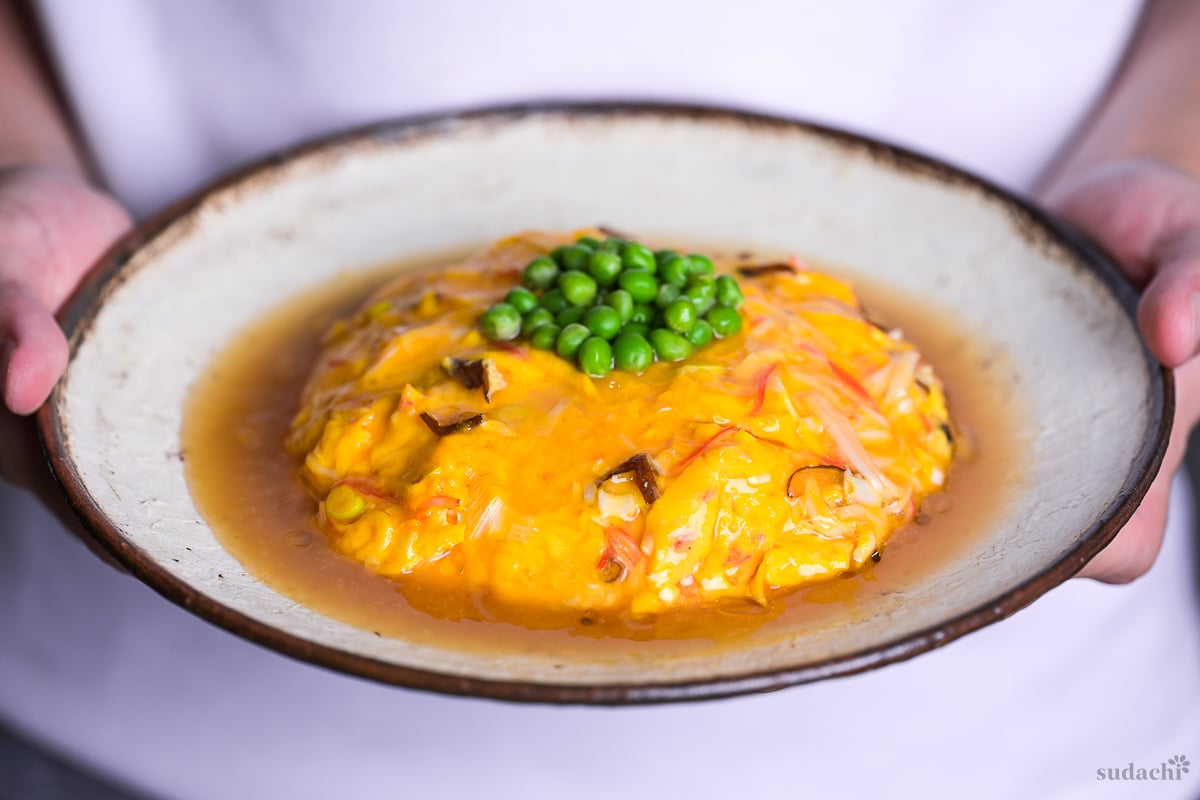 Yuto Omura holding up tenshinhan (Japanese crab omelette with rice) topped with green peas on a textured cream plate with brown rim