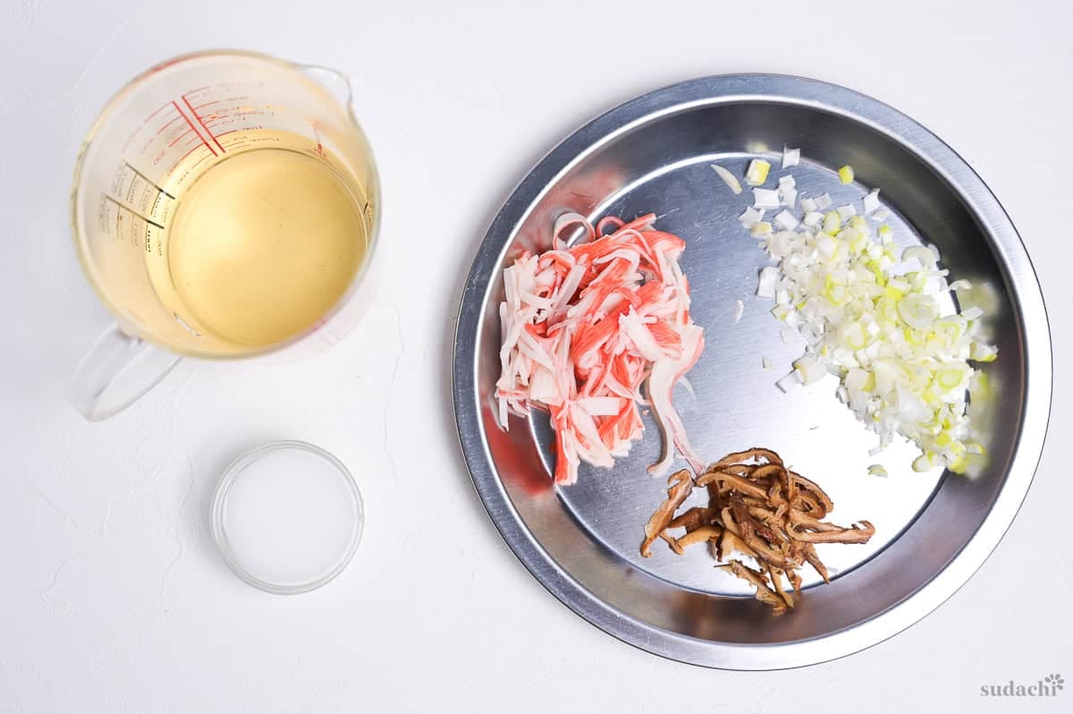 shiitake dashi in a jug next to a slurry made with potato starch and cold water in a small glass bowl, and a silver prep plate with shredded imitation crab sticks, shredded shiitake mushrooms and finely diced Japanese leek (negi)