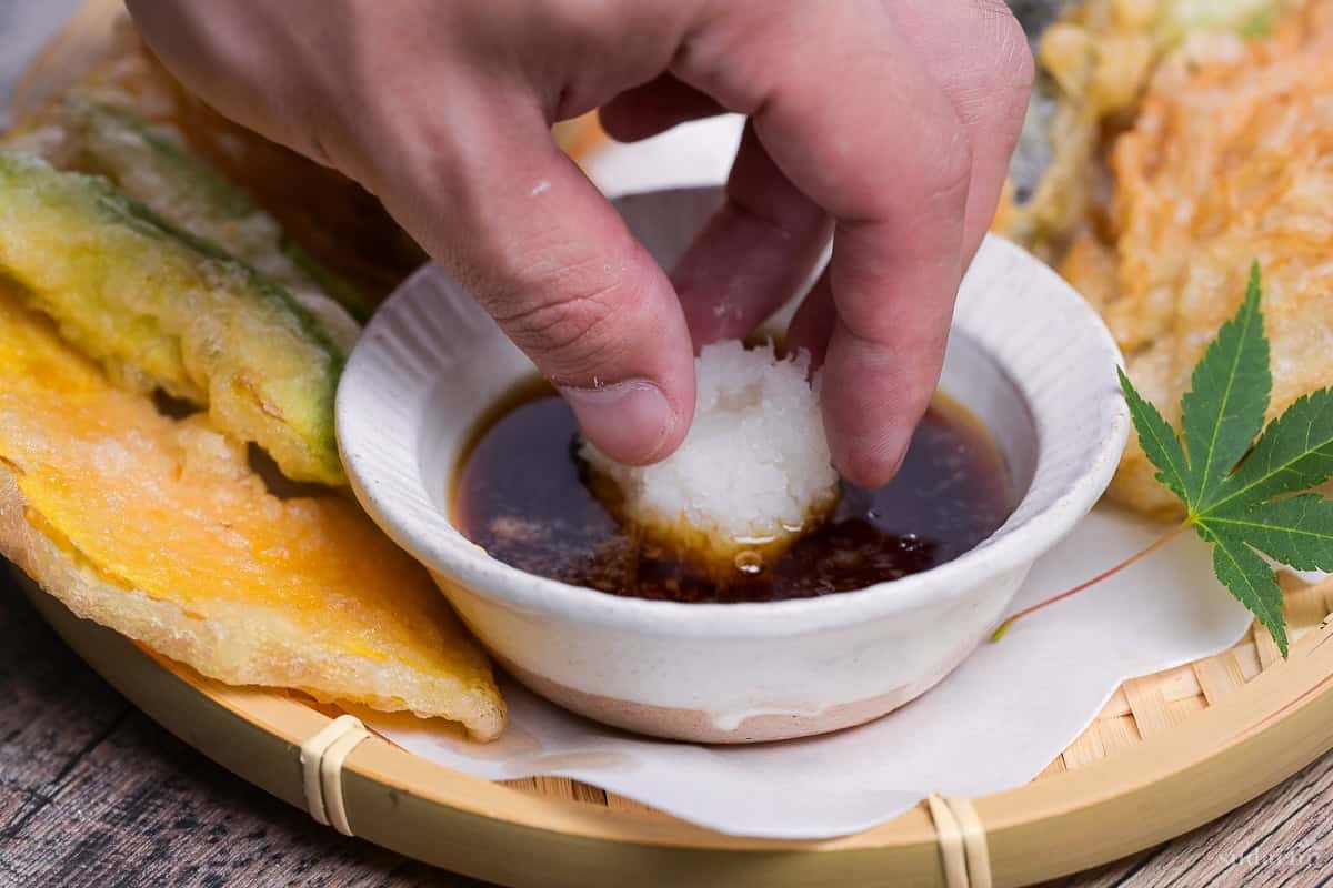 a hand placing a mound of grated daikon in a dish of homemade tsuyu tempura dipping sauce