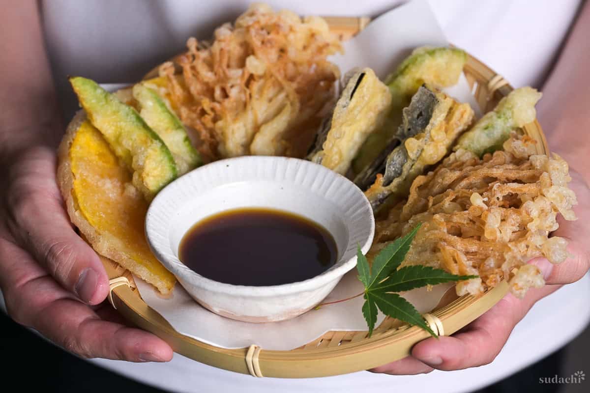 Yuto Omura holding up homemade tentsuyu (tempura dipping sauce) in a white bowl surrounded by vegetable tempura on a round bamboo tray