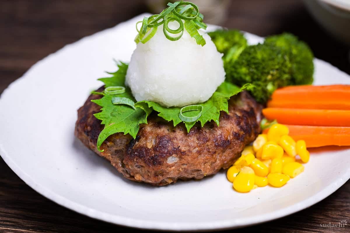 homemade wafu hamburg topped with a perilla leaf, grated daikon radish and chopped green onions next to steamed broccoli, carrots and sweet corn on a white plate