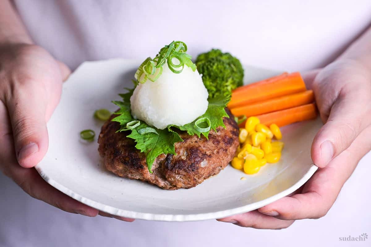 Yuto Omura holding homemade wafu hamburg topped with a perilla leaf, grated daikon radish and chopped green onions next to steamed broccoli, carrots and sweet corn on a white plate