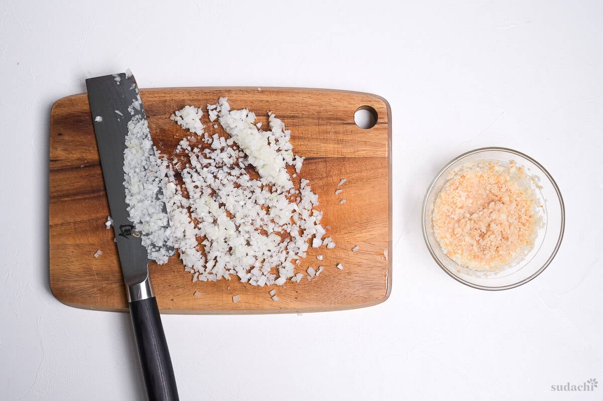 finely diced onion on a wooden cutting board with knife, next to a bowl of panko mixed with sake to make a panade.