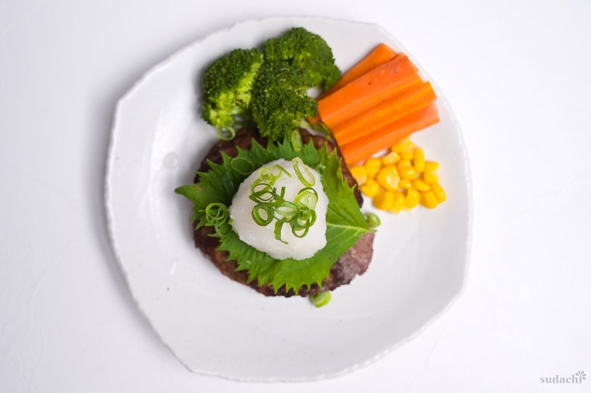 Wafu hamburg topped with a perilla leaf, grated daikon and finely chopped green onions next to steamed broccoli, carrots and sweet corn on a white plate