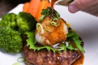 Pouring homemade sauce over wafu hamburg topped with a perilla leaf, grated daikon radish and chopped green onions next to steamed broccoli, carrots and sweet corn on a white plate