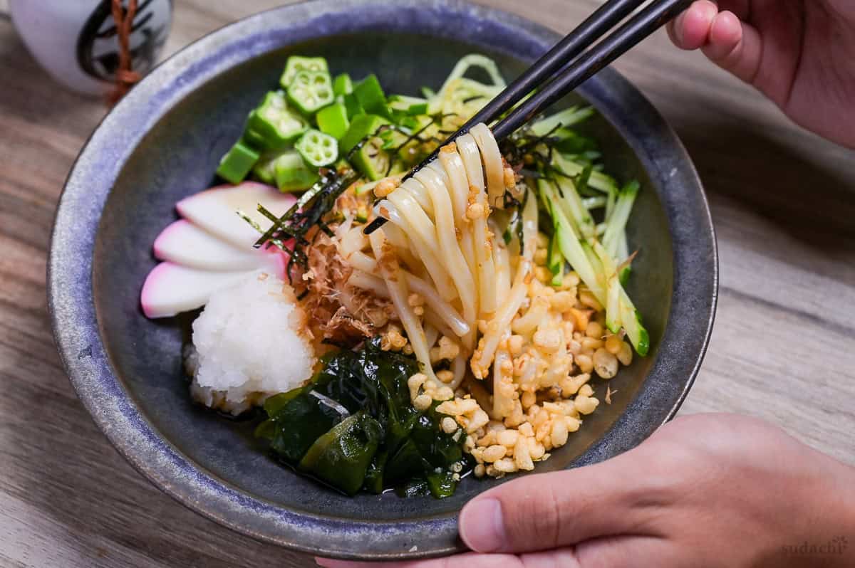 Hiyashi tanuki udon topped with wakame, grated daikon radish, kamaboko slices, okra, cucumber and tempura bits in a dark blue-gray dish with chopsticks lifting the udon