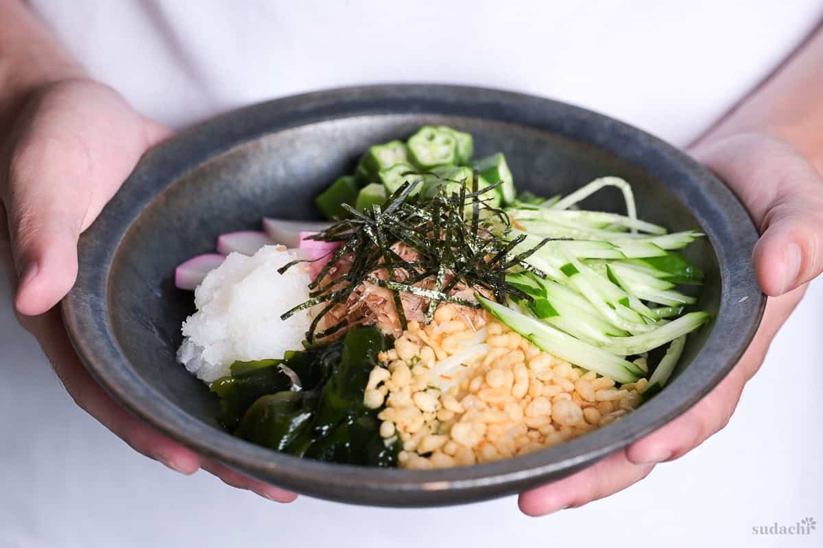 Yuto Omura holding up Hiyashi tanuki udon topped with wakame, grated daikon radish, kamaboko slices, okra, cucumber and tempura bits in a dark blue-gray dish
