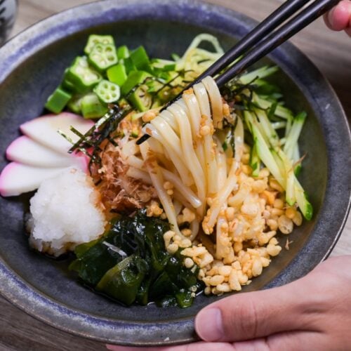 Hiyashi tanuki udon topped with wakame, grated daikon radish, kamaboko slices, okra, cucumber and tempura bits in a dark blue-gray dish