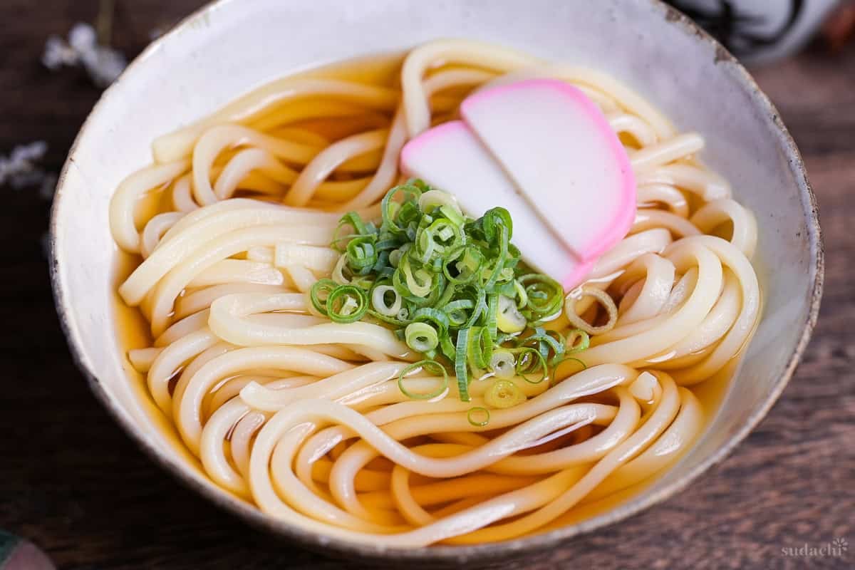 homemade kake udon noodle soup topped with chopped green onions (negi) and two slices of kamaboko Japanese fish cakes in an off-white ceramic bowl with brown rim