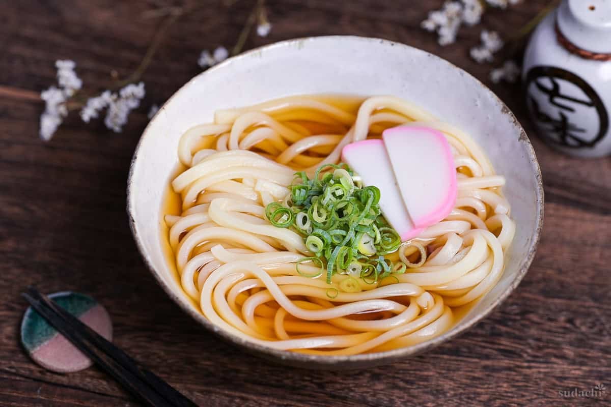 homemade kake udon noodle soup topped with chopped green onions (negi) and two slices of kamaboko Japanese fish cakes in an off-white ceramic bowl with brown rim