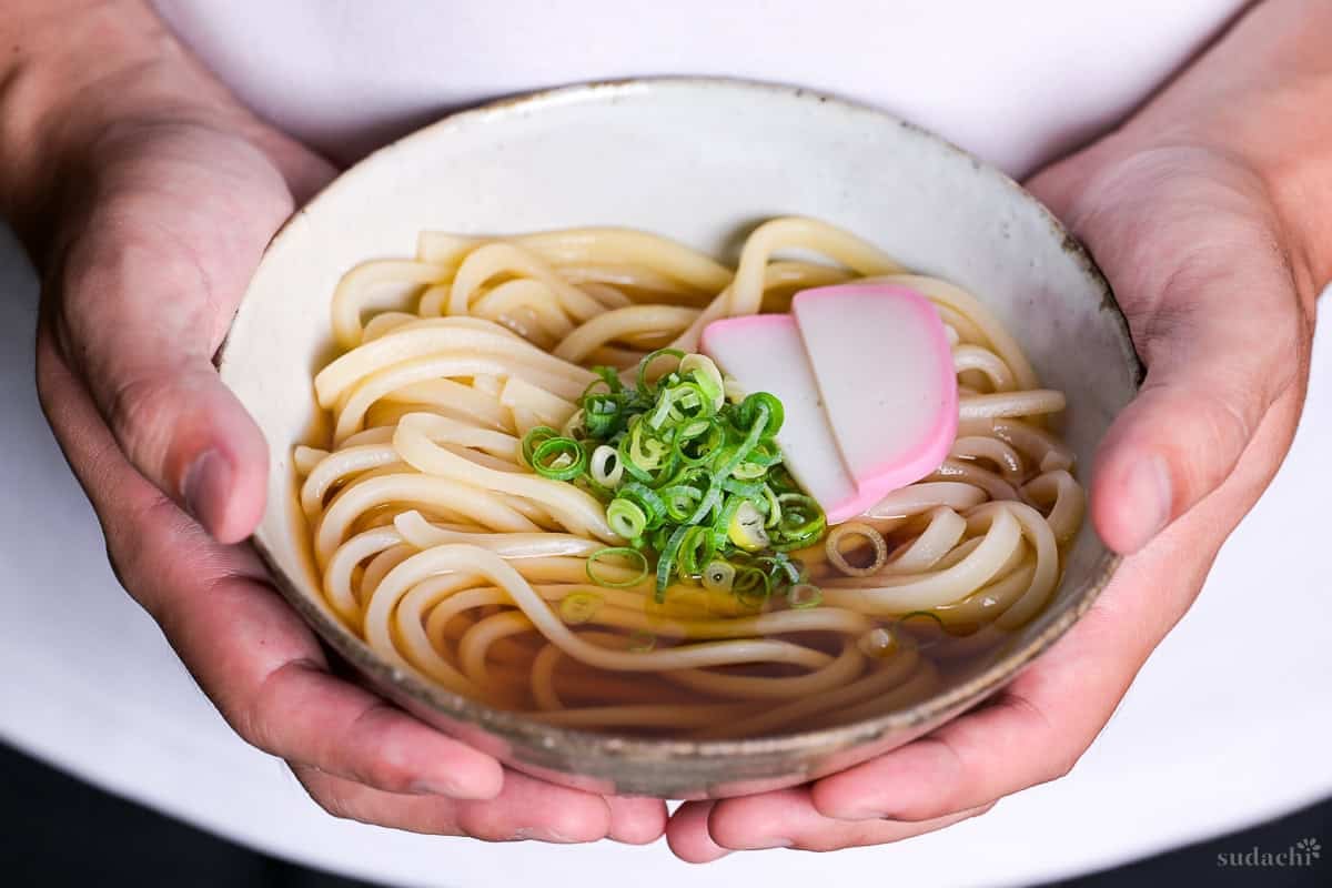 Yuto Omura holding up a bowl of homemade kake udon noodle soup topped with chopped green onions (negi) and two slices of kamaboko Japanese fish cakes