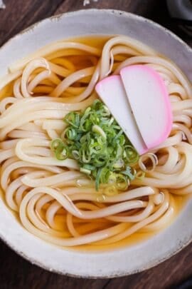 homemade kake udon noodle soup topped with chopped green onions (negi) and two slices of kamaboko Japanese fish cakes in an off-white ceramic bowl with brown rim