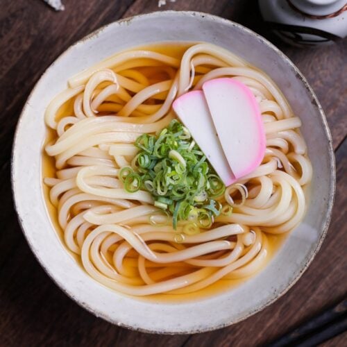 homemade kake udon noodle soup topped with chopped green onions (negi) and two slices of kamaboko Japanese fish cakes in an off-white ceramic bowl with brown rim