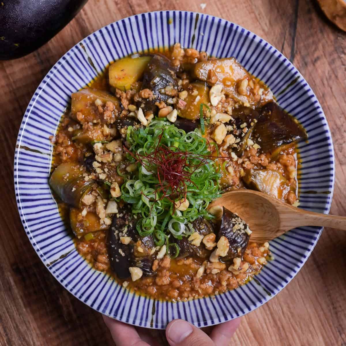 Plant-based mabo nasu (mapo eggplant) topped with chopped green onion and chili threads served in a white dish with blue striped edge with wooden spoon taking a scoop thumbnail square