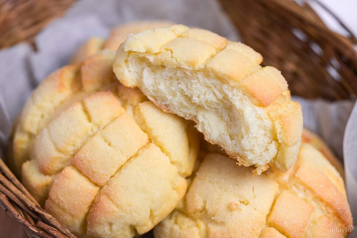 Melon pan torn in half to reveal pillowy bread inside placed on top of whole melon pans in a wicker basket