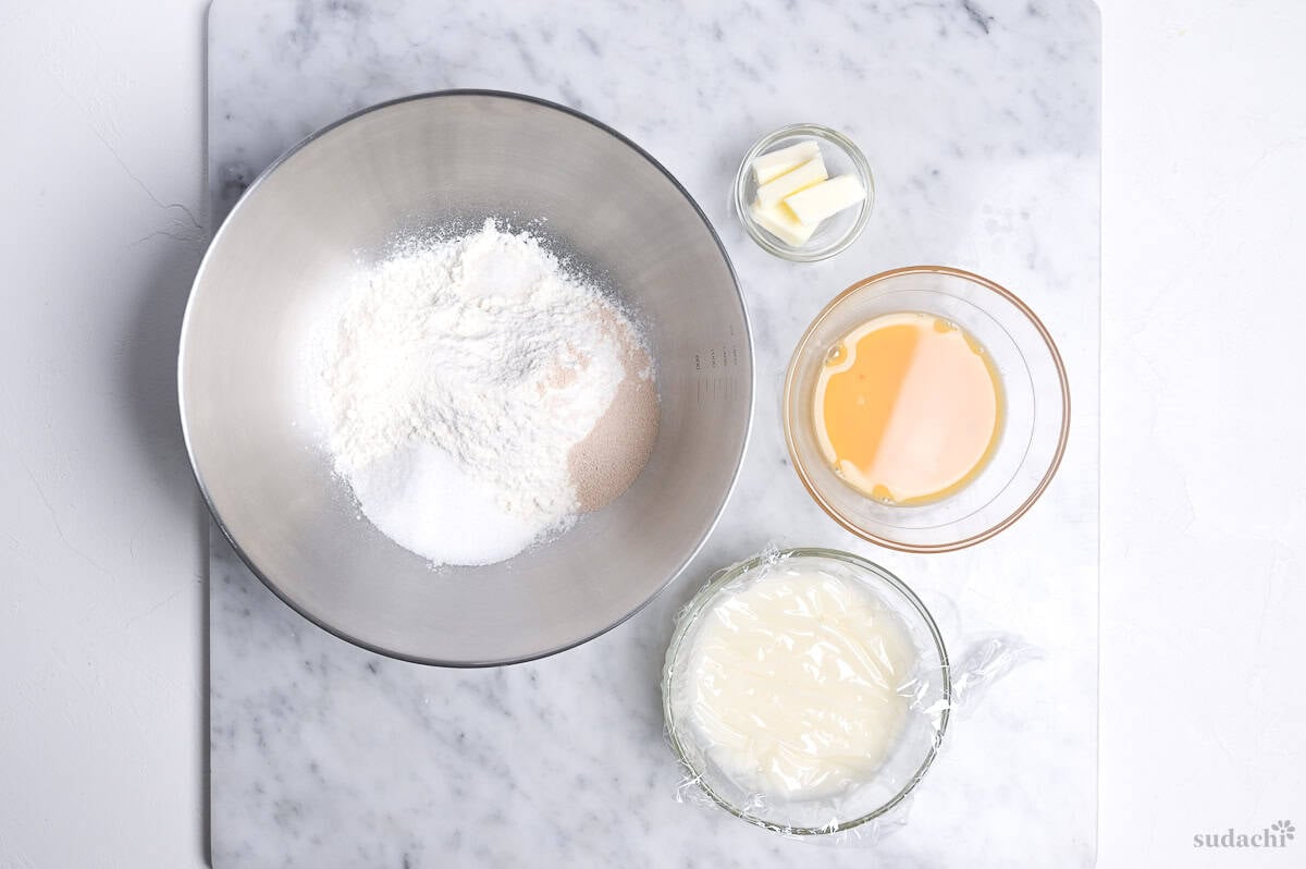 elements of melon pan bread on a kneading board. Dry ingredients in a steel mixing bowl, butter measured in a small glass bowl, egg and honey mixed in a bowl, tangzhong in a bowl.