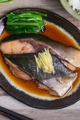 Sakana no nitsuke made with yellowtail topped with julienned ginger and served with blanched greens on a oval plate with brown rim thumbnail