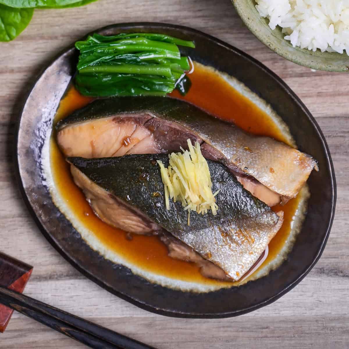 Sakana no nitsuke made with yellowtail topped with julienned ginger and served with blanched greens on a oval plate with brown rim thumbnail