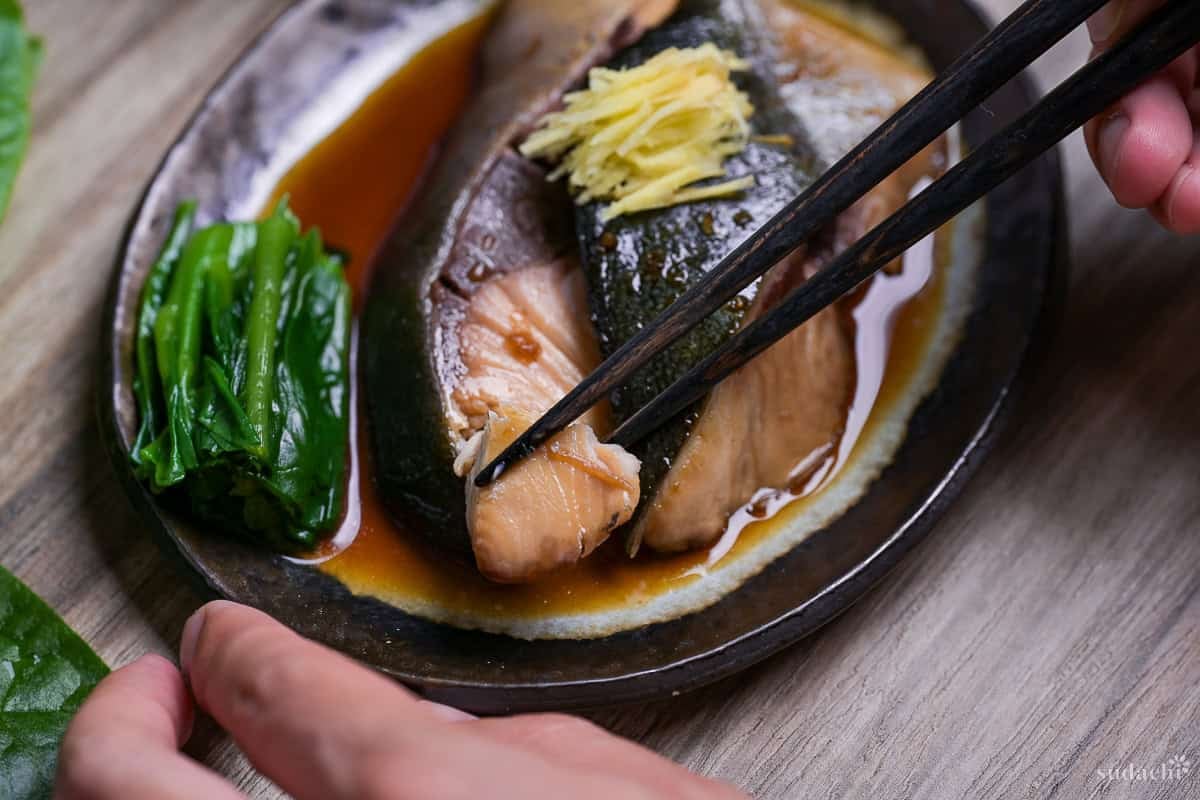 Sakana no nitsuke made with yellowtail topped with julienned ginger and served with blanched greens on a oval plate with brown rim with black chopsticks lifting a bit of fish