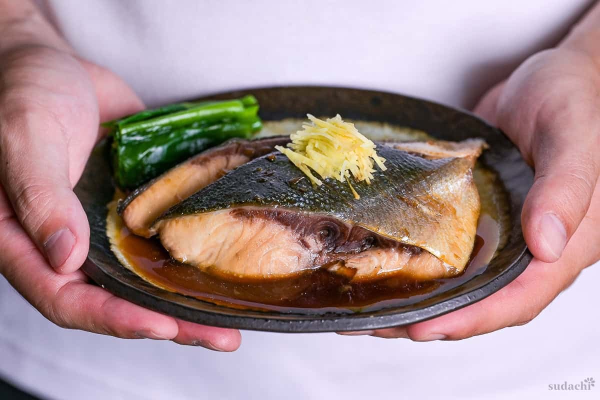 Yuto Omura holding Sakana no nitsuke made with yellowtail topped with julienned ginger and served with blanched greens on a oval plate with brown rim