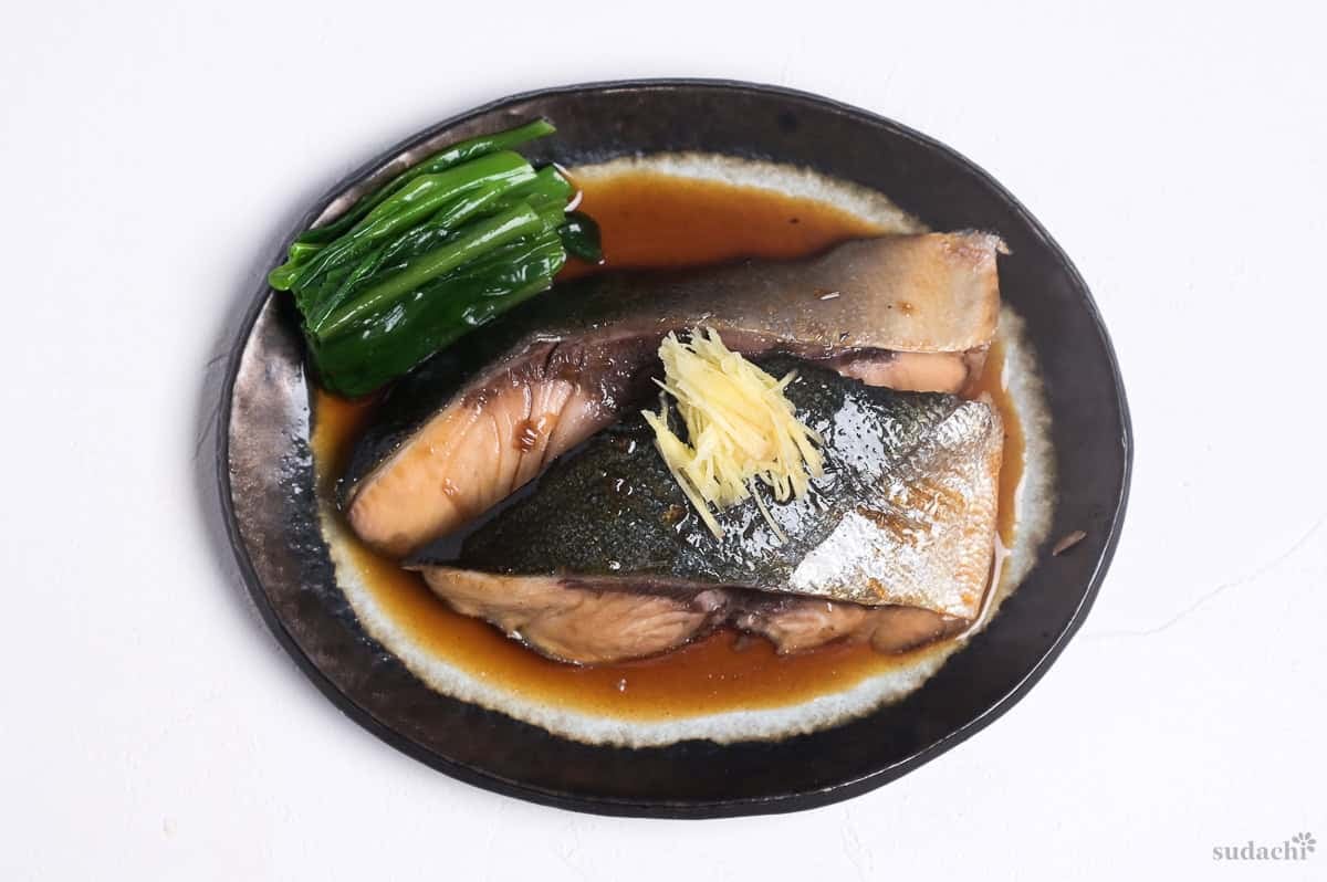 sakana no nitsuke made with yellowtail fillets topped with julienned ginger and served with blanched greens on an oval plate with brown rim.