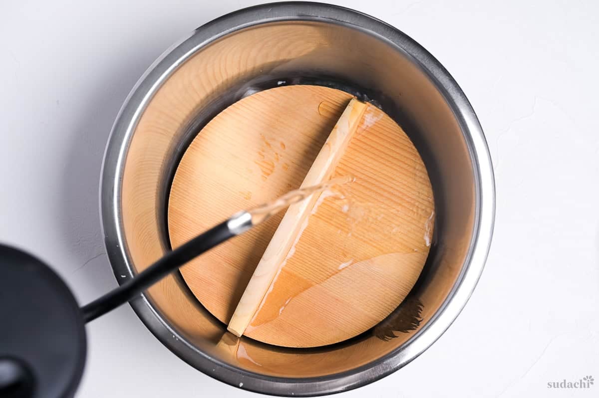 fish fillet covered with a wooden drop lid in a steel mixing bowl with freshly boiled water being poured over the top