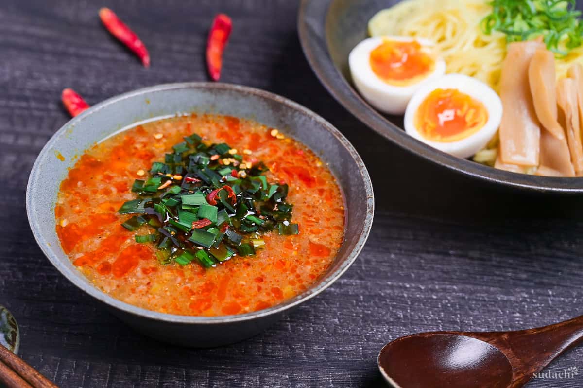 Spicy tsukemen dipping ramen in a blue bowl next to a plate with ramen noodles, soft boiled eggs and menma, and dried chili peppers in the background