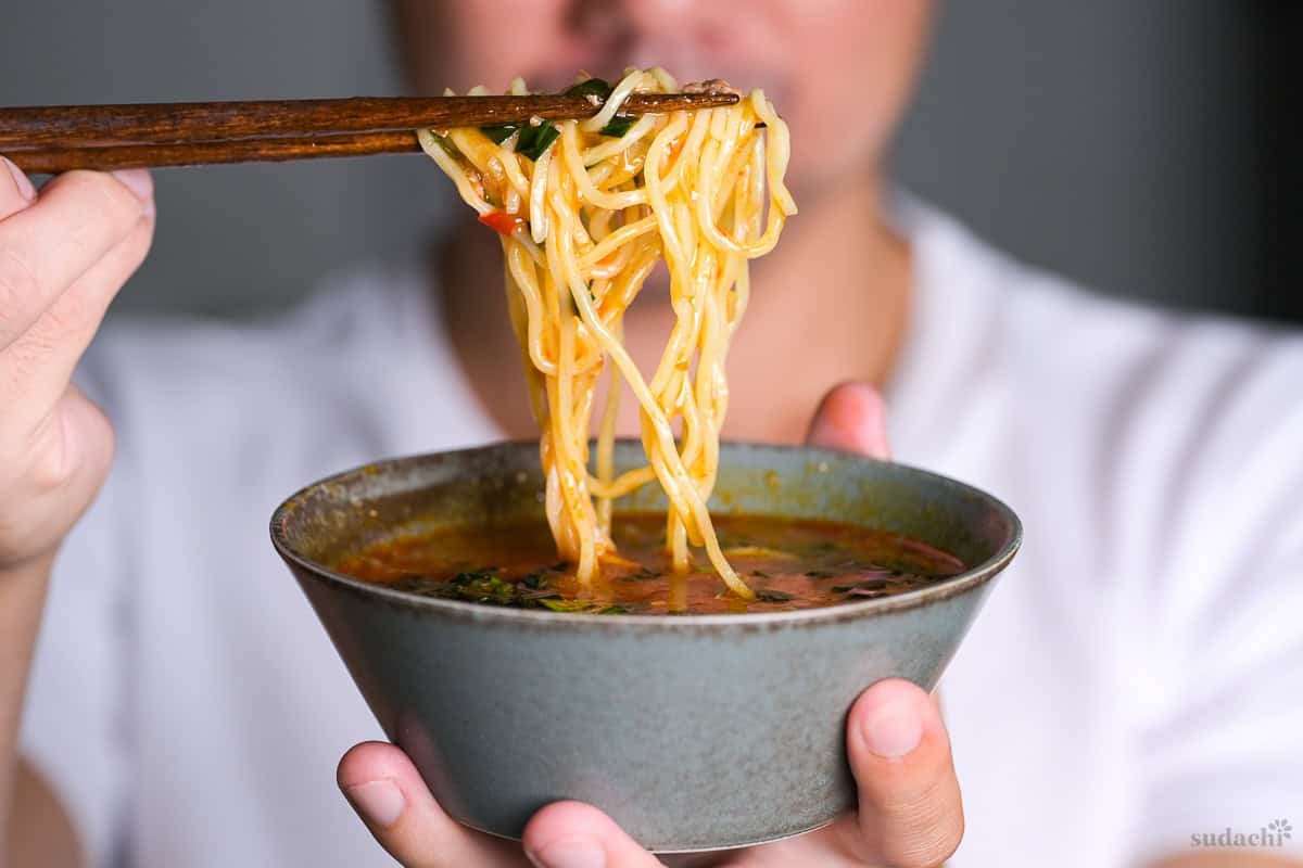 Yuto Omura holding up a bowl of spicy tsukemen dipping soup with ramen noodles held with wooden chopsticks above it