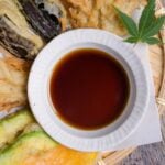 Homemade tentsuyu (tempura dipping sauce) in a white bowl surrounded by vegetable tempura served on a bamboo tray top down