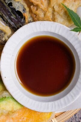 Homemade tentsuyu (tempura dipping sauce) in a white bowl surrounded by vegetable tempura served on a bamboo tray top down