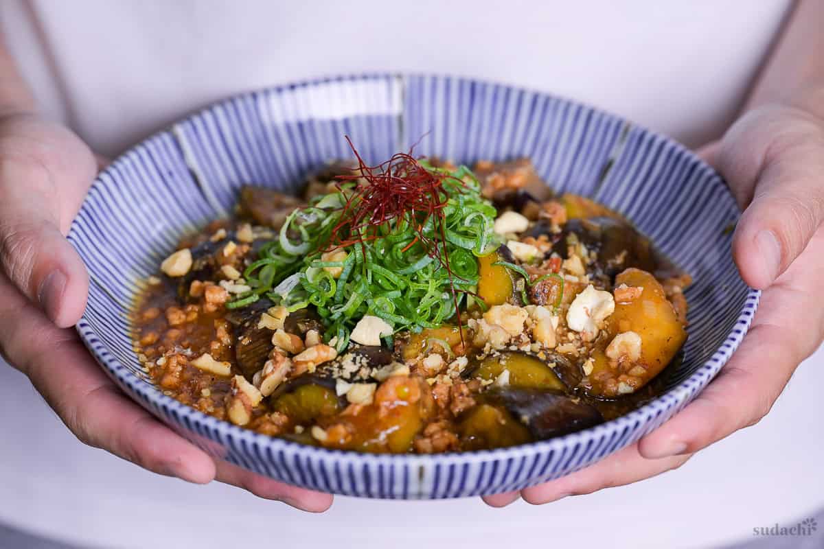 Yuto Omura holding plant-based mabo nasu (mapo eggplant) topped with chopped green onion and chili threads served in a white dish with blue striped edge