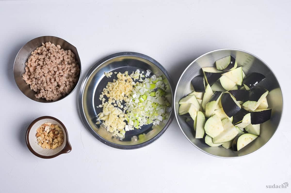 Mis en place for plant-based mabo nasu: tofu mince in a bowl, crushed cashew nuts in a mini mortar, finely chopped garlic, ginger and leek on a steel plate, and roughly cut Japanese eggplant in a steel mixing bowl.