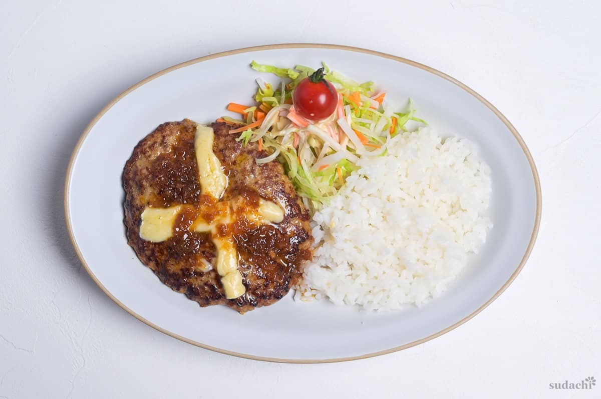 Bikkuri Donkey Inspired hamburg steak topped with a cross of melted cheese, next to white rice and salad served on a white oval plate on a white background
