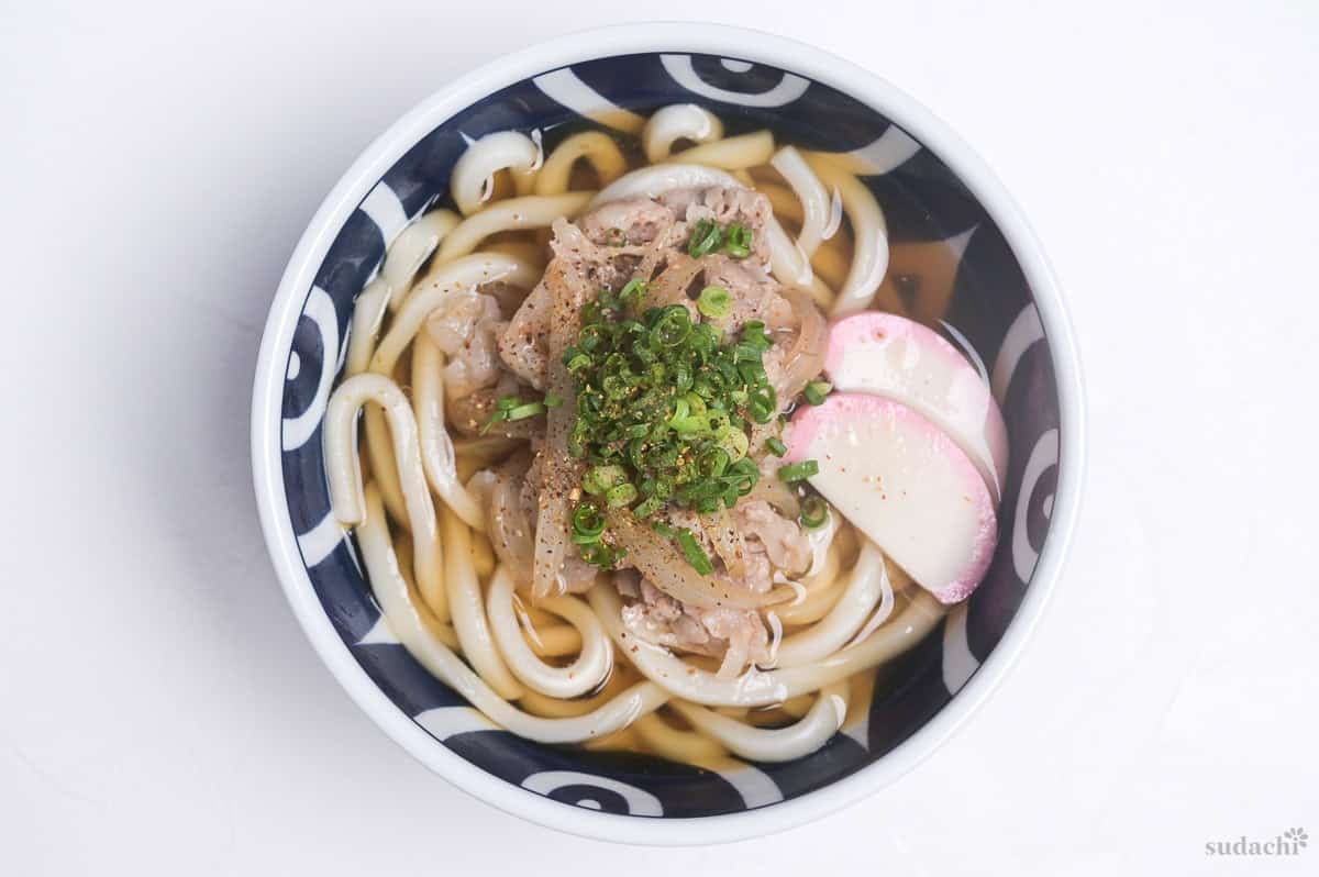 Pork udon noodle soup topped with chopped green onions, shichimi togarashi and kamaboko fish cakes in a white and blue bowl