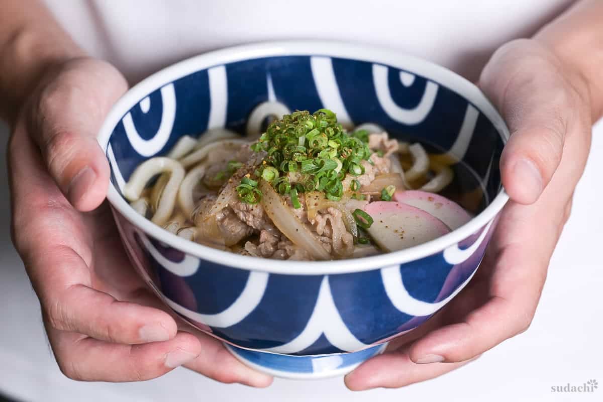 Yuto Omura holding pork udon noodle soup topped with kamaboko fishcakes, chopped green onion and shichimi togarashi in a blue bowl with white swirl design