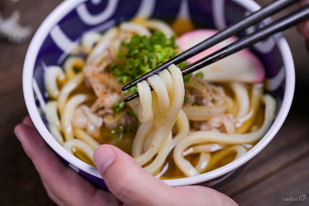 pork udon noodle soup topped with kamaboko fishcakes, chopped green onion and shichimi togarashi in a blue bowl with white swirl design with black chopsticks holding up the noodles