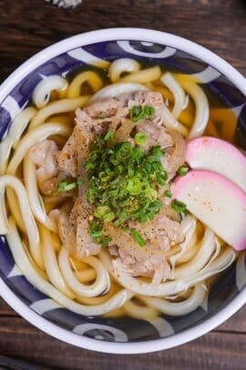 pork udon noodle soup topped with kamaboko fishcakes, chopped green onion and shichimi togarashi in a blue bowl with white swirl design top down view