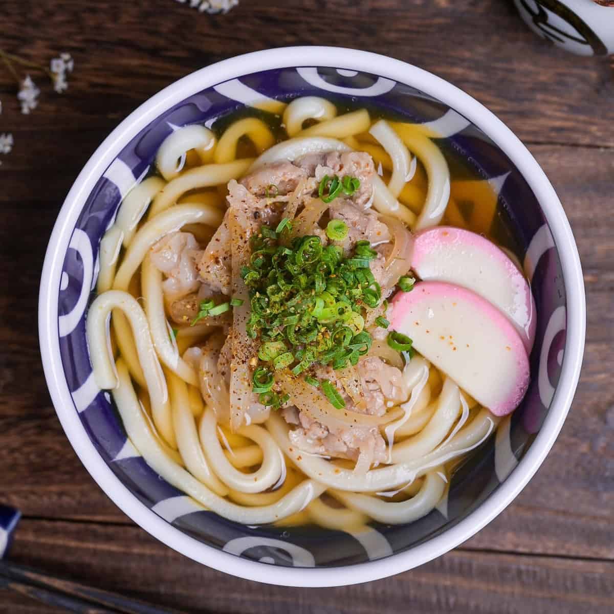 pork udon noodle soup topped with kamaboko fishcakes, chopped green onion and shichimi togarashi in a blue bowl with white swirl design top down view